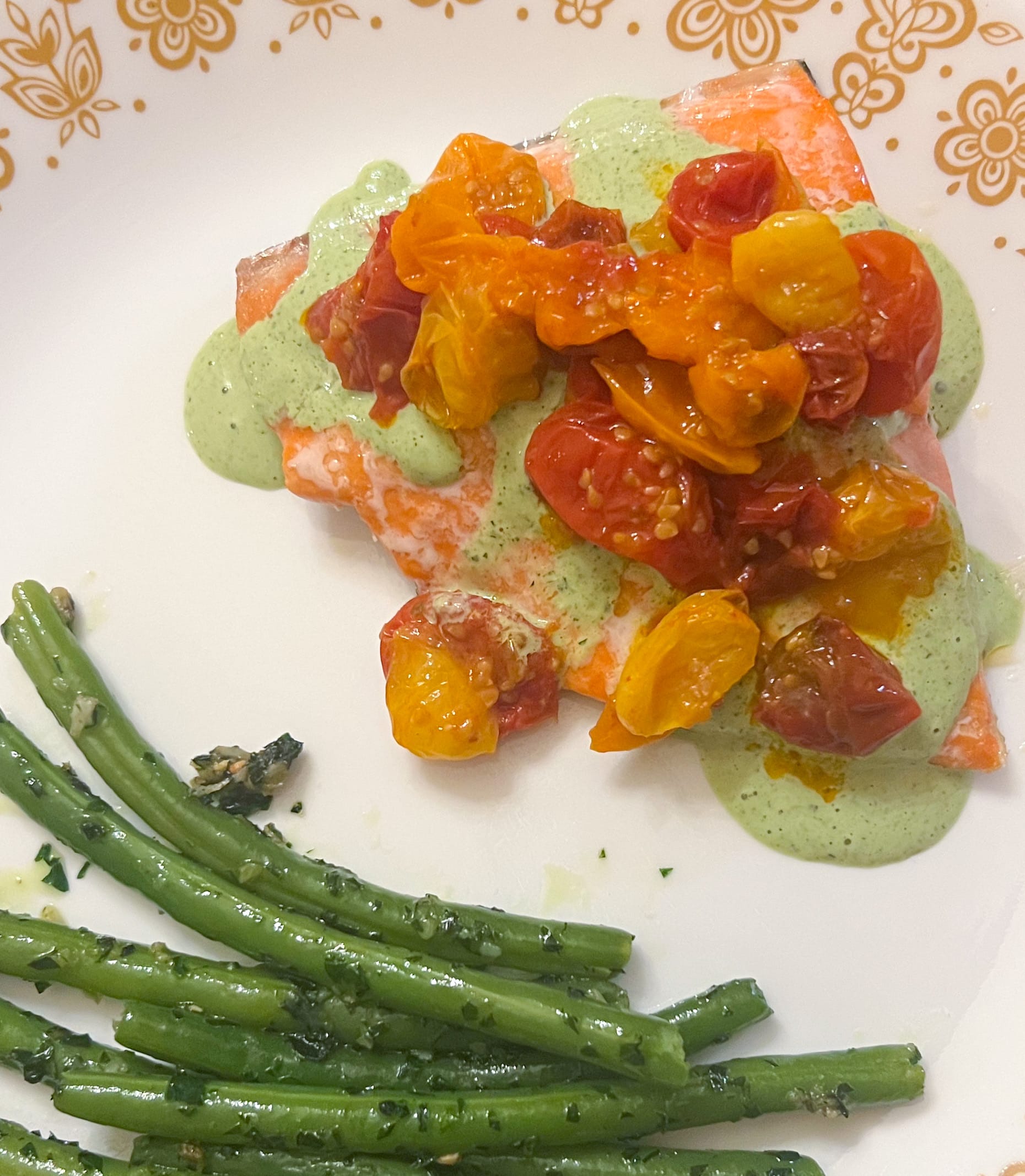 A piece of salmon is topped with Green Goddess dressing and yellow, orange, and red cherry tomatoes. There are garlicky green beans on the side.