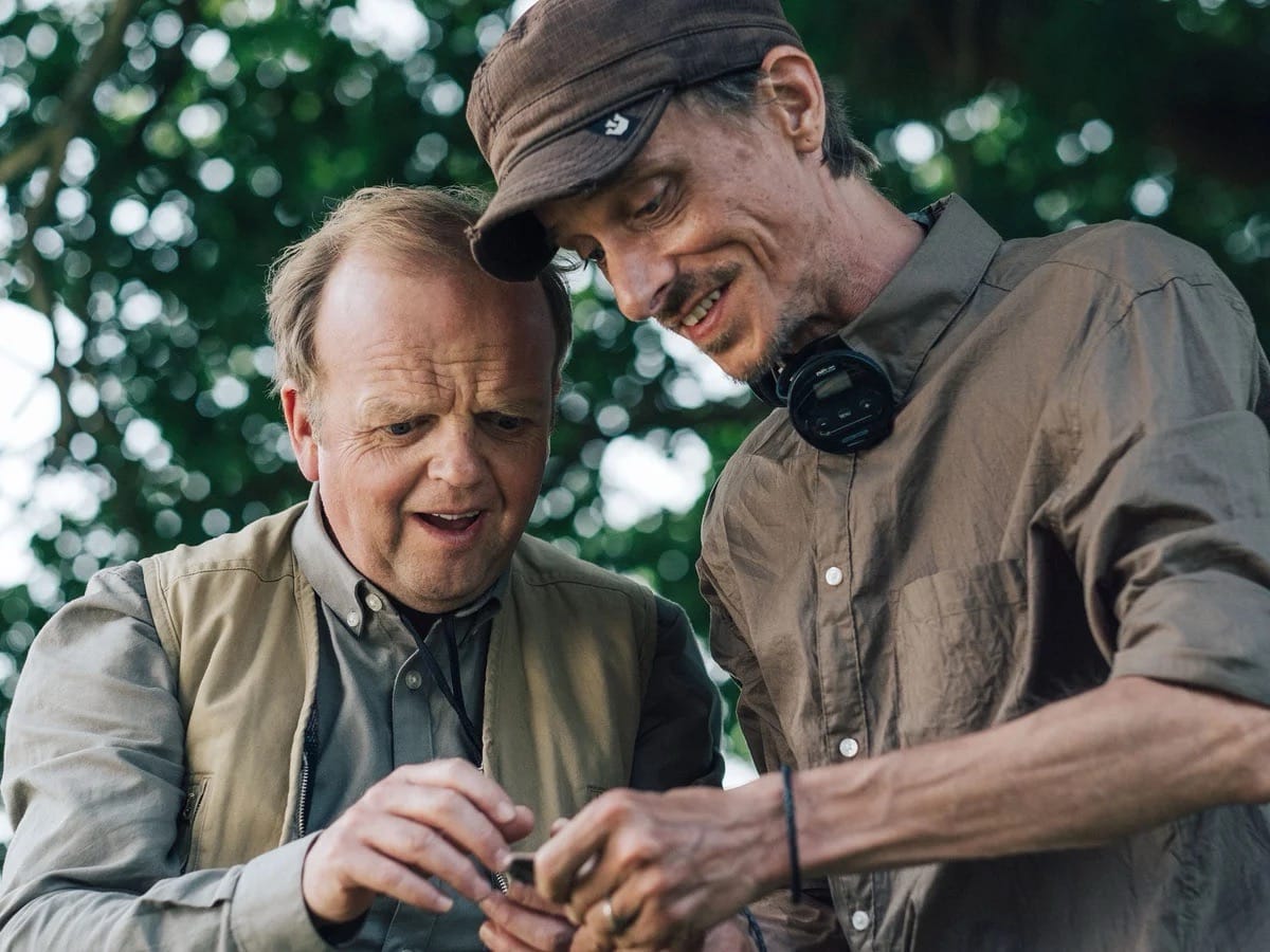 Two men stand together under a tree, one is showing a small object to the other, who looks excited to see it.