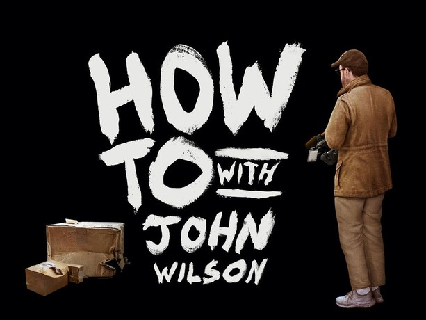 A man with a camera has his back to us. White pained letters say "How To with John Wilson". Busted up cardboard boxes are at the left.