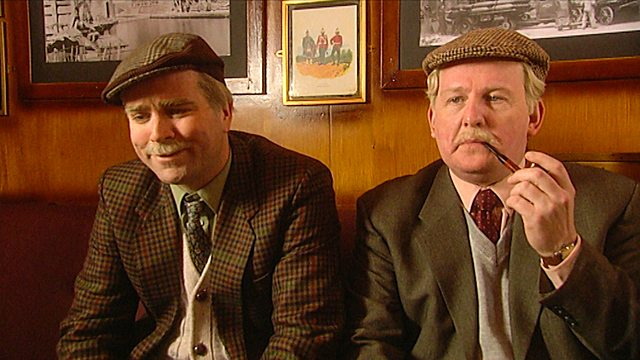 Two elderly white men dressed in tweed jackets and caps sit near the wall of a pub.