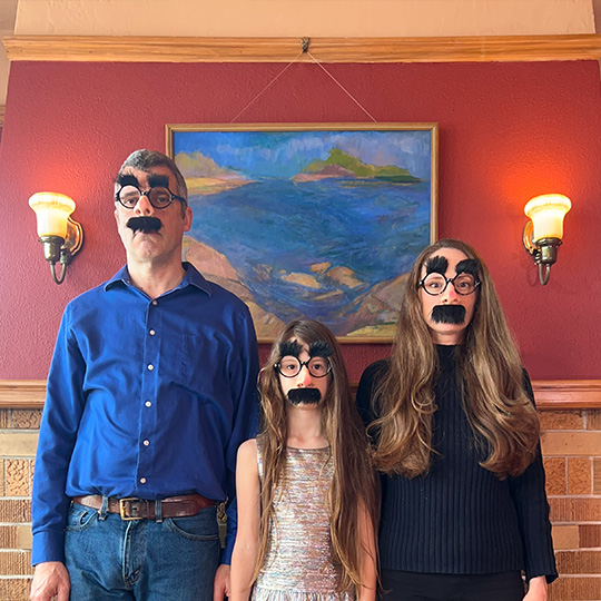 A family of three, including a preteen daughter, looking at the camera seriously while wearing Groucho Marx glasses.