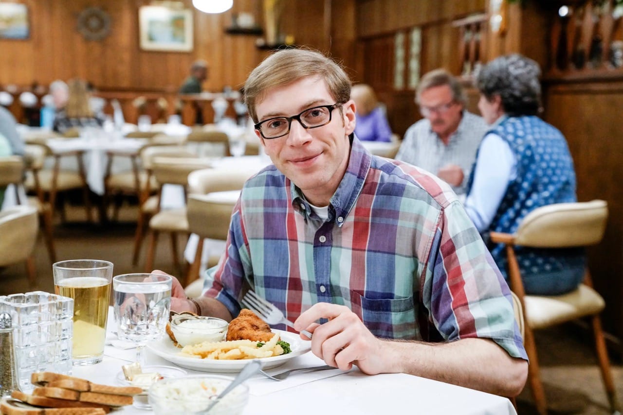 A bespectacled man is pausing from eating his breakfast in a family restaurant, to look at you warmly.