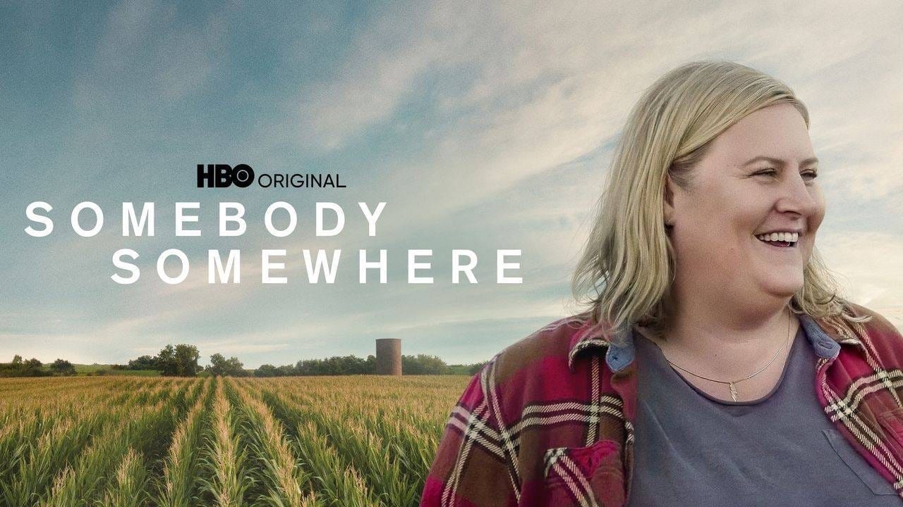 A cornfield stretches off in the distance. In to foreground, a blond woman in a red flannel with an open smile looks away.