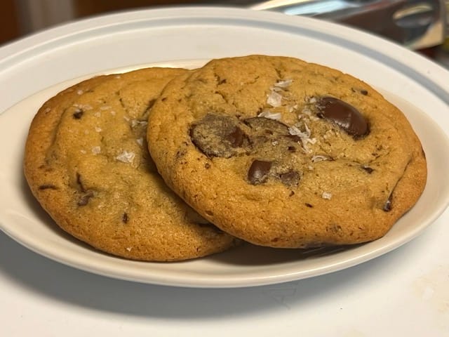 Two chocolate chip cookies, topped with large flakes of salt, on plate.