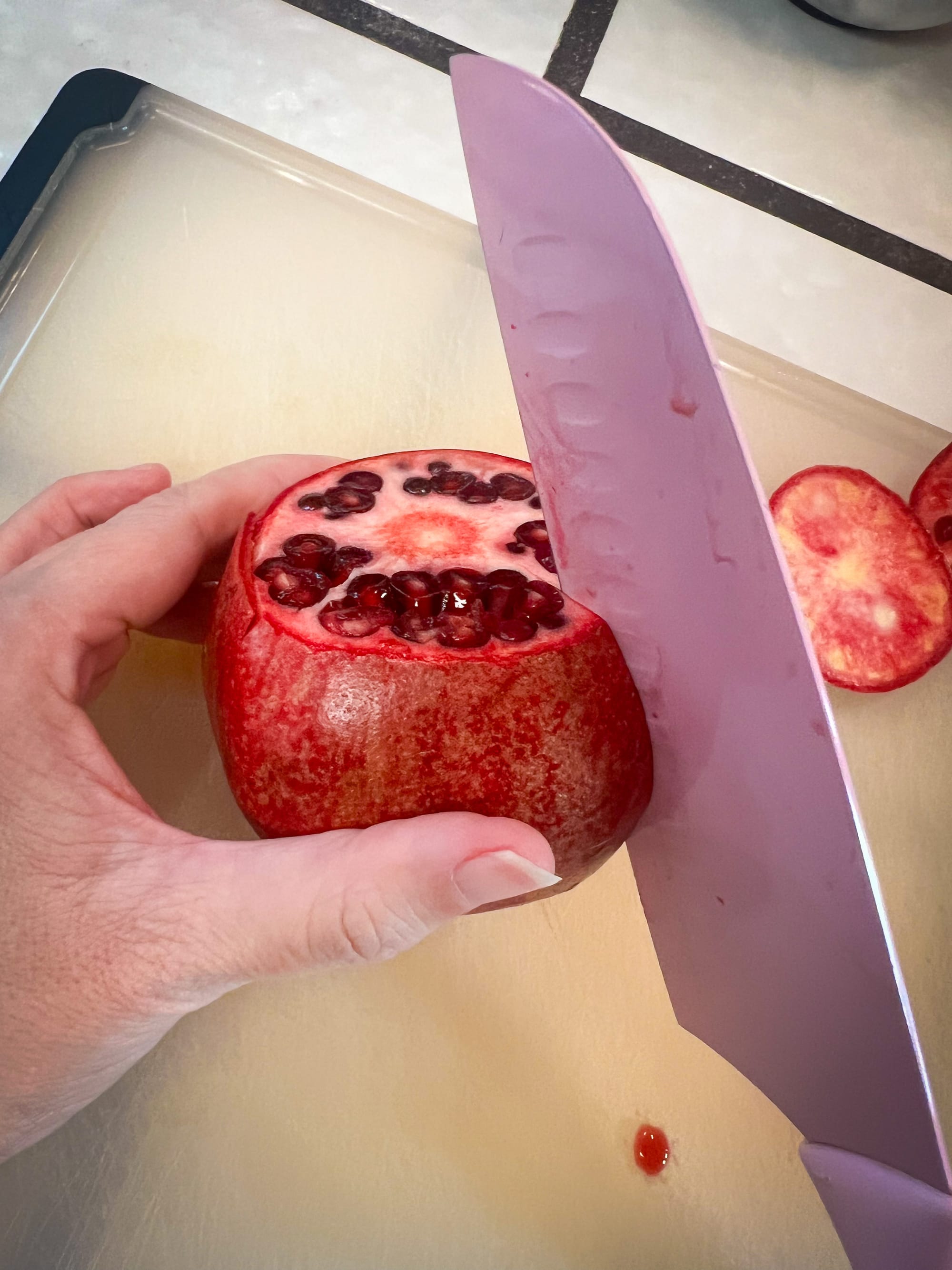 Left hand is holding a pomegranate with its top cut off, while a knife cuts into the outer rind vertically, top to bottom. The knife is only a couple millimeters into the fruit.