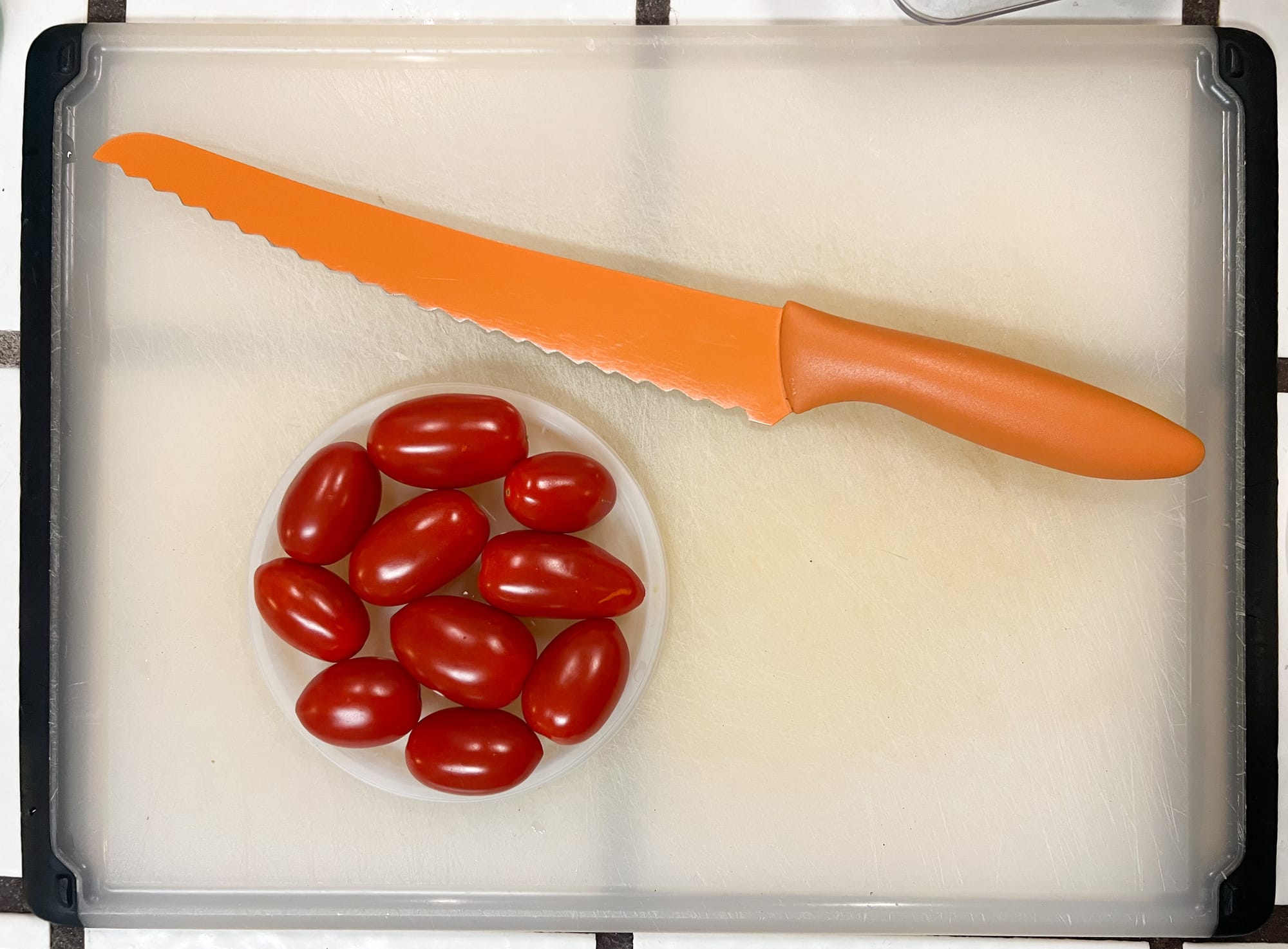 Ten red grape tomatoes are nestled in a round plastic lid, alongside a long, orange, serrated bread knife.