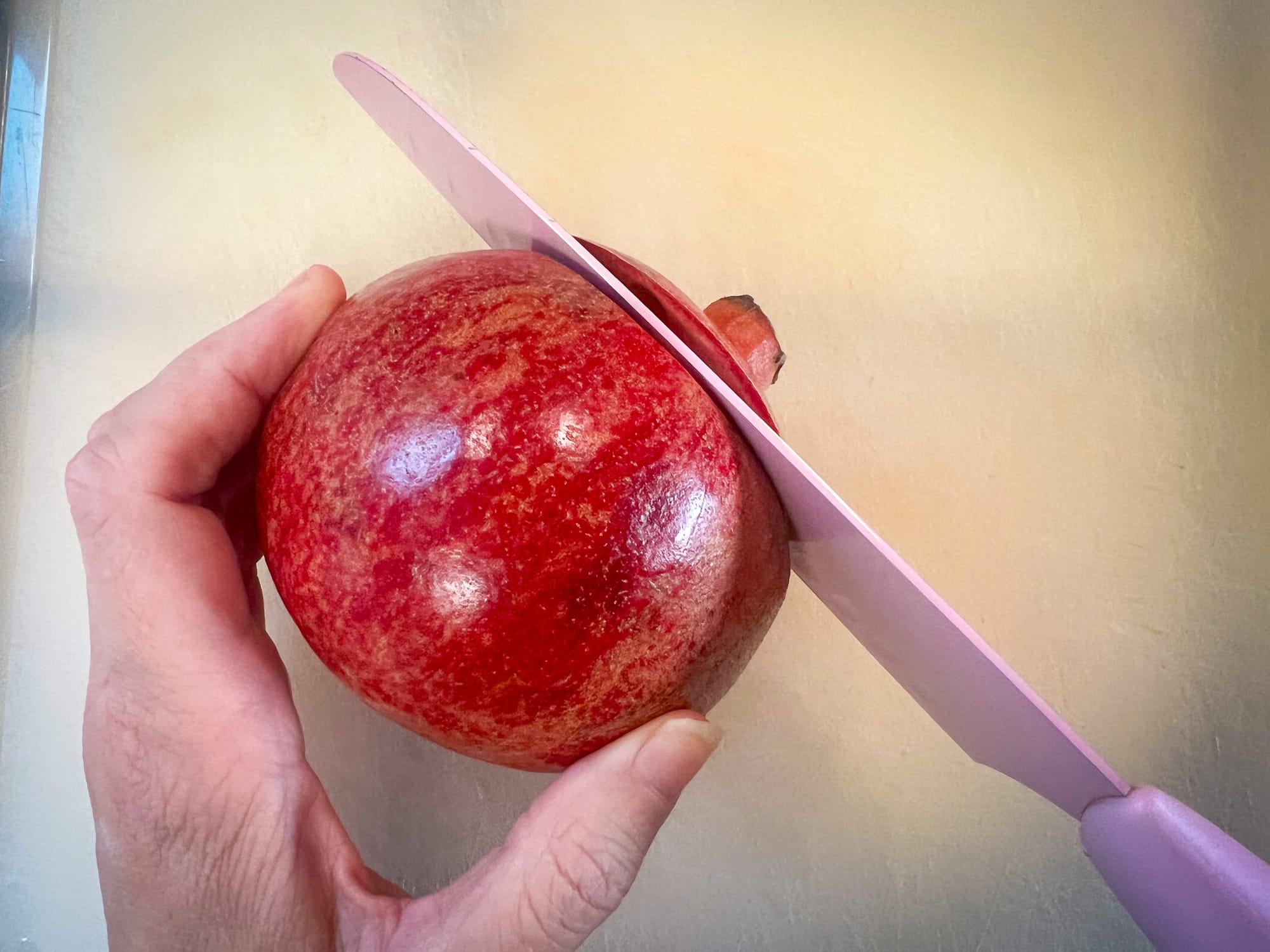 A pomegranate is held with a left hand while the right makes a cut in the top. It's very close to the top, about a quarter-inch.