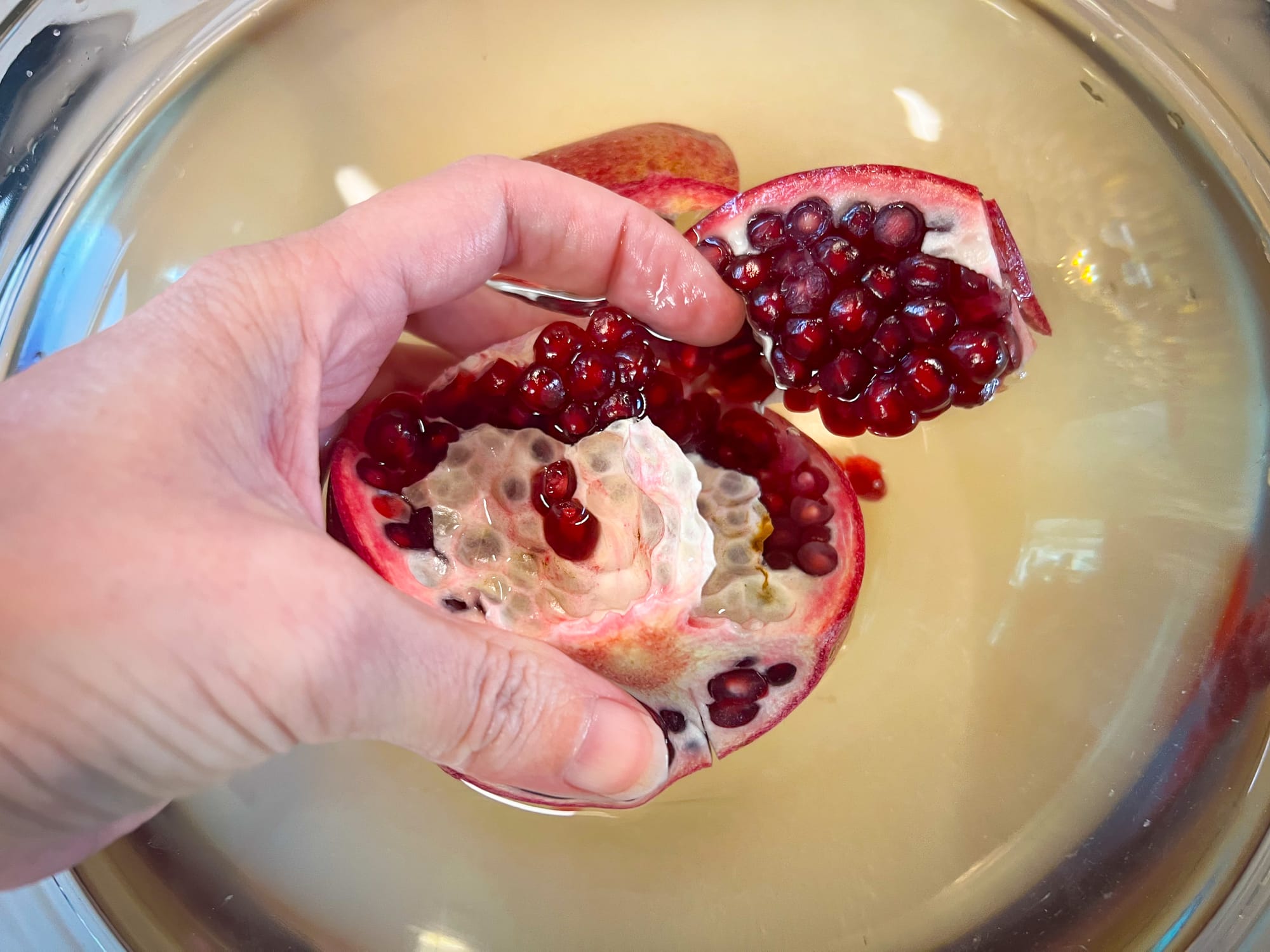 A hand grips the submerged pomegranate, and is pulling a big chunk apart. Lots of pomegranate seeds are now visible, along with thin white membrane.