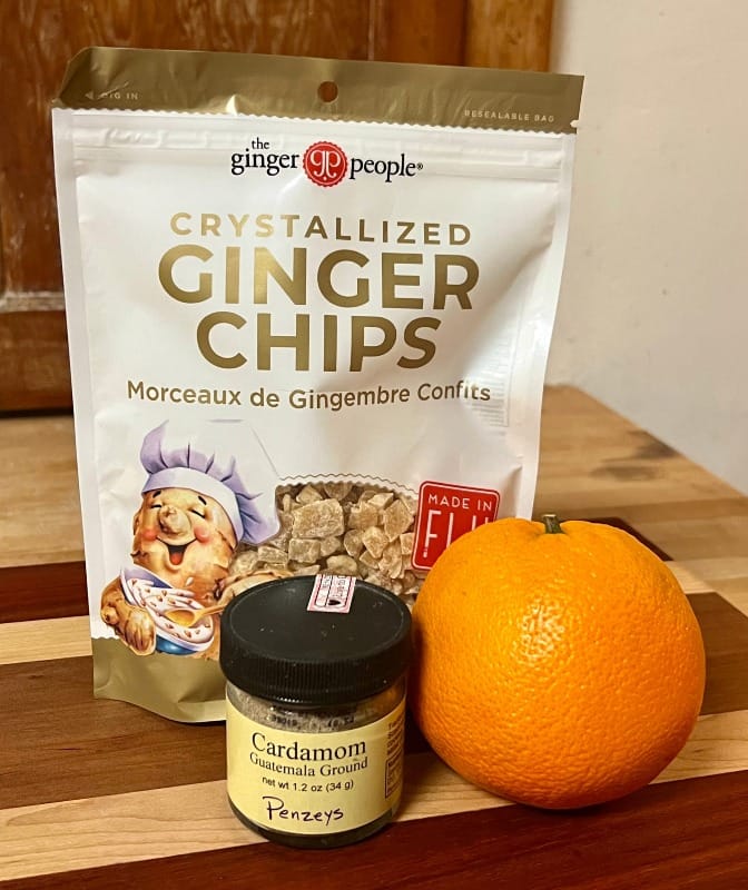 A bag of ginger chips, a small jar of ground cardamom, and an orange.