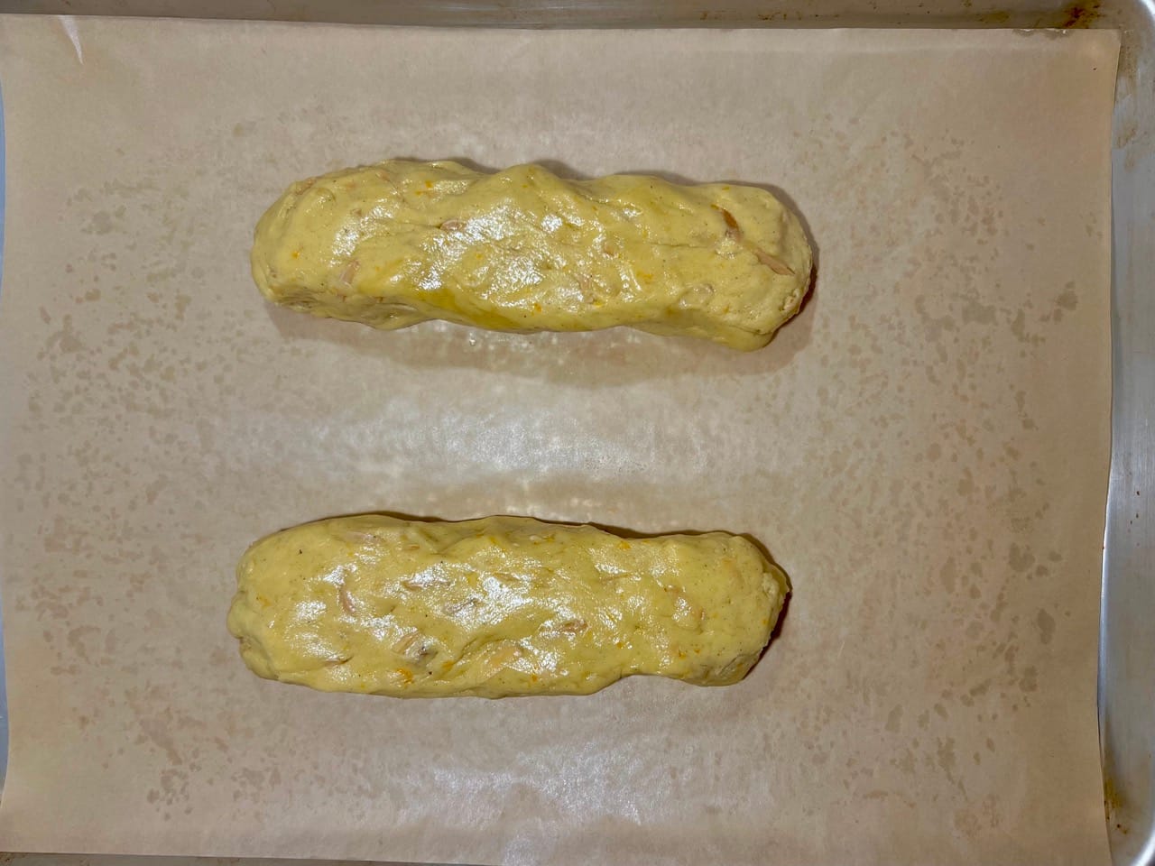 Two unbaked, unshaped logs of biscotti dough are side by side on a parchment-lined baking sheet.