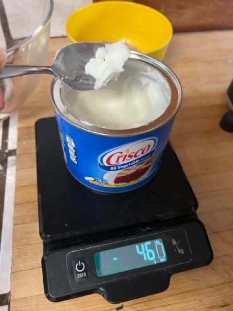 A container of Crisco shortening is on a digital scale, which reads -46 grams. A spoon above the container has removed some shortening.