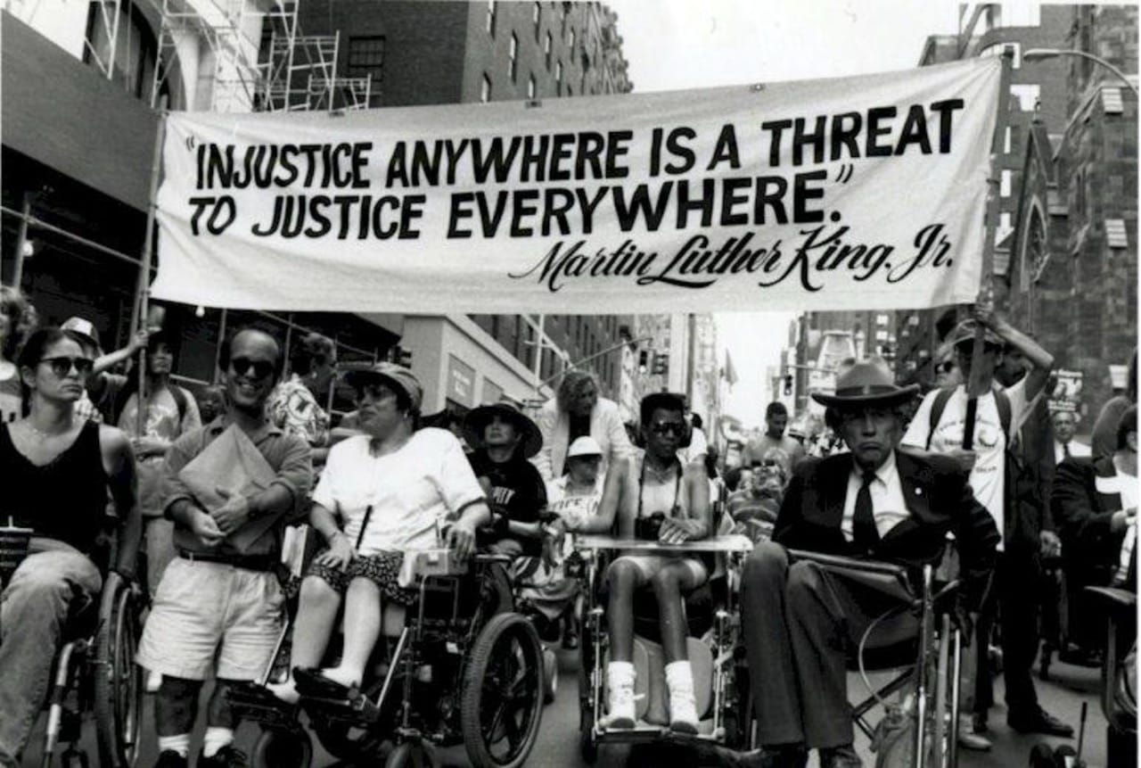 Disabled people march below a banner saying "Injustice anywhere is a threat to justice everywhere" by MLK