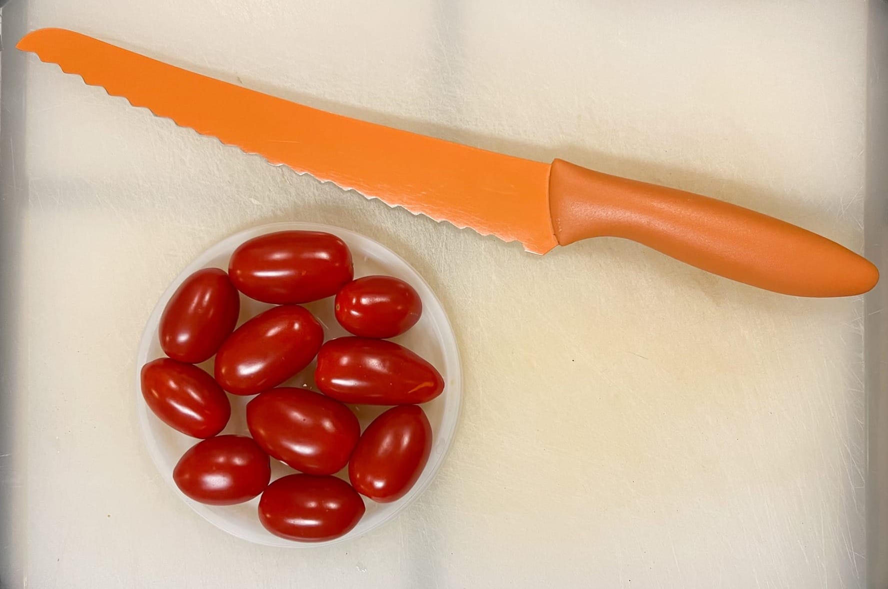 Ten red grape tomatoes are nestled in a round plastic lid, alongside a long, orange, serrated bread knife.