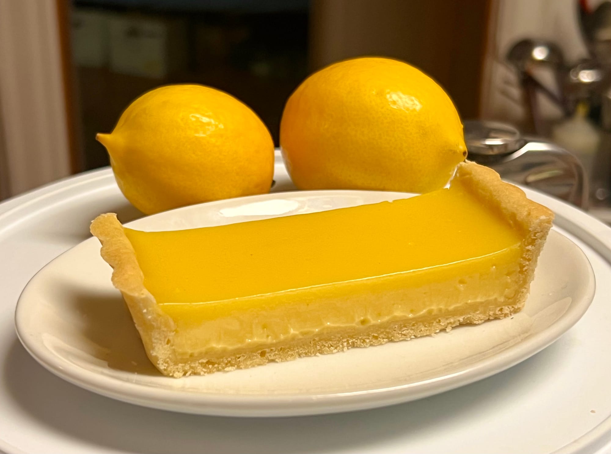 A rectangular slice of bright yellow lemon tart, with two Meyer lemons behind.