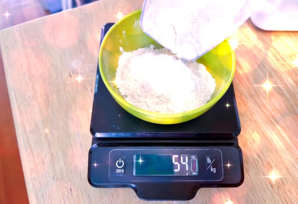 Flour is being poured into a bowl on a digital kitchen scale, which reads 54g.