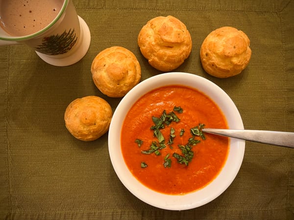 A bowl of creamy red tomato soup, topped with basil, with four gougères on the side.