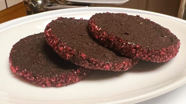 Three round cookies on a plate. They're a dark chocolatey brown, with jewel-like pink sugar sprinkles around the outer edge.