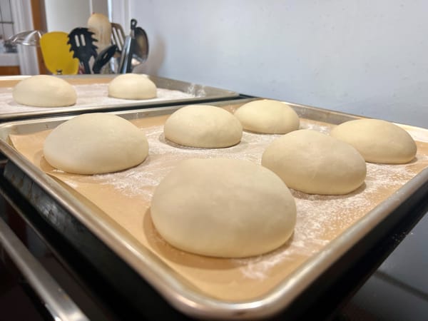 Eight round balls of unbaked pizza dough are arranged on two floured baking sheets.