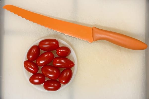 Ten red grape tomatoes are nestled in a round plastic lid, alongside a long, orange, serrated bread knife.