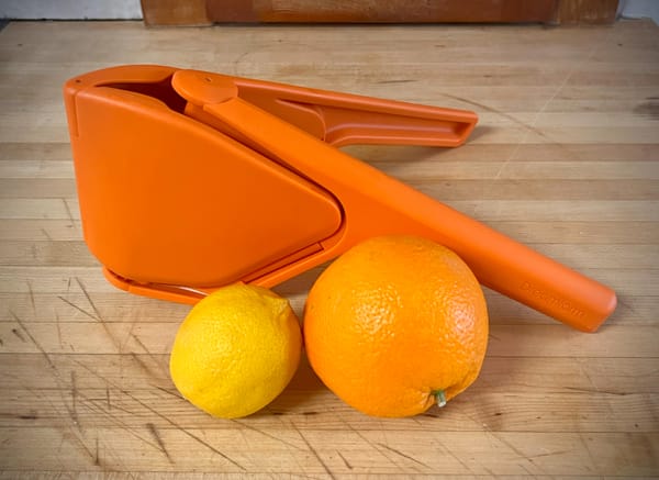 An orange plastic tool is on a counter next to an orange and a lemon. The tool is shaped like a V, with two handles.